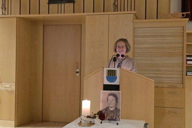 Frau an einem Rednerpult aus Holz, davor SW-Porträt einer Frau, daneben brennende Kerze