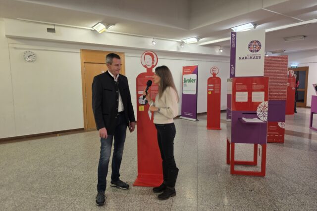 Frau mit Mikrofon mit Mann in Anzug vor einer Symbolfigur